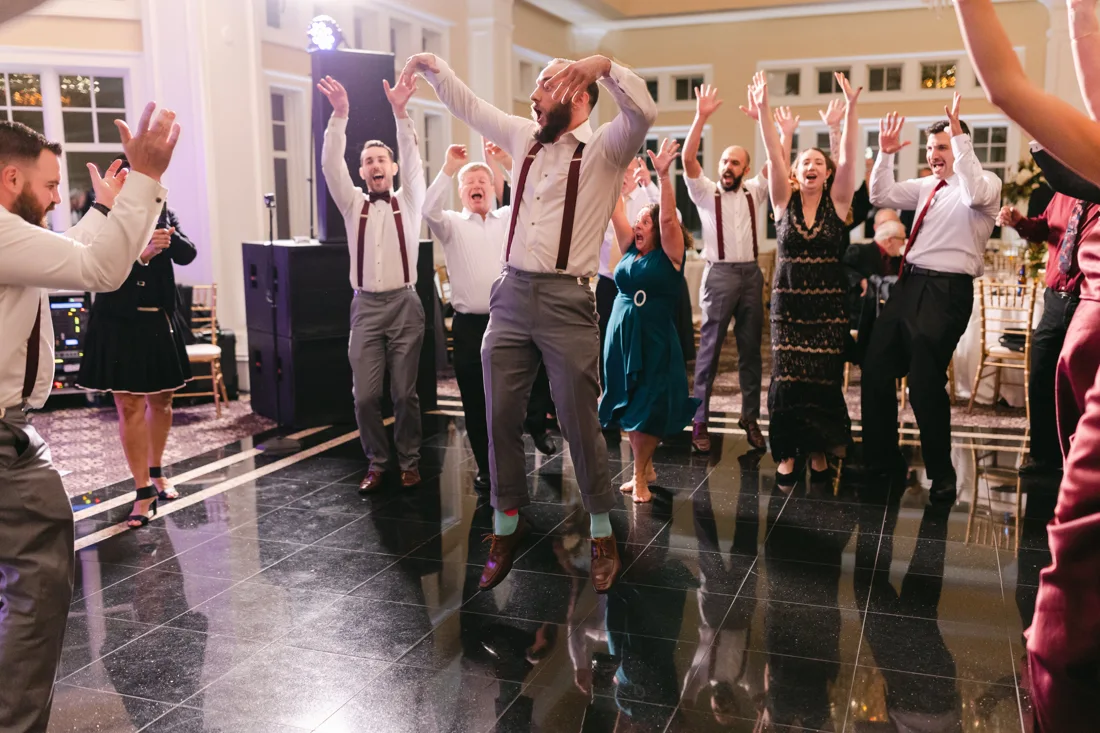 Groomsmen and guests jumping with arms raised on a packed reception dance floor