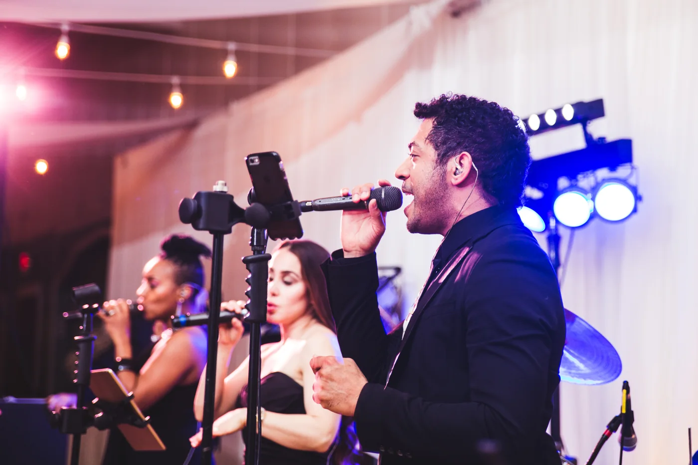 Three Live Wire vocalists performing together under warm string lights